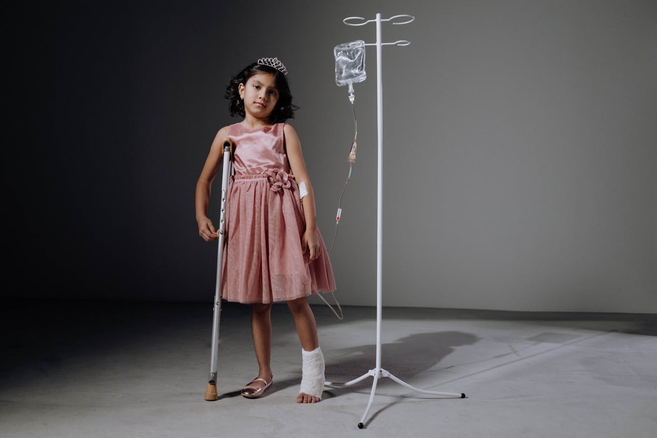 Injured girl in pink dress using crutches next to an IV stand in a studio setting.